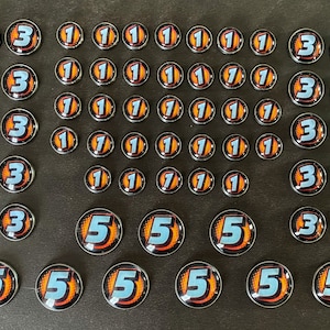 Puede incluir: Una colecci&oacute;n de fichas de juego redondas de color plateado con n&uacute;meros naranja y azul. Los n&uacute;meros incluyen 1, 3 y 5.
