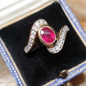May include: A gold ring with a large oval ruby centre stone. The ring features a wave design with small, round, clear stones set along the curves. The ring is displayed in a blue jewellery box with a black velvet interior.
