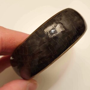May include: A black bangle bracelet with a textured, reptile-like pattern. The bracelet is made of metal with a gold-toned finish.