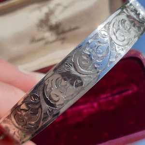 May include: A silver bangle with detailed floral and scroll engraving. The polished metal reflects light, showcasing the intricate patterns. The bracelet is presented against a burgundy velvet background.