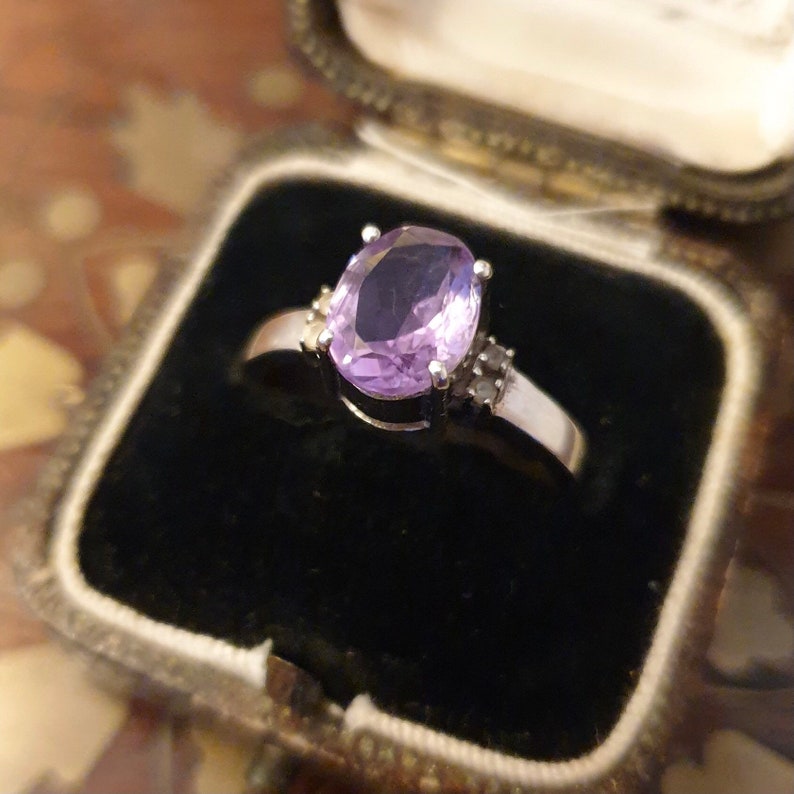 May include: A silver ring with a large oval amethyst gemstone and smaller diamond accents. The ring is displayed in a black velvet ring box.