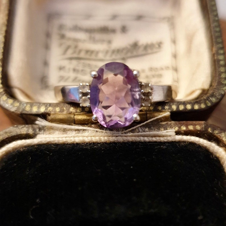 May include: A silver ring with a large oval amethyst gemstone and smaller diamond accents. The ring is in a velvet-lined jewelry box.
