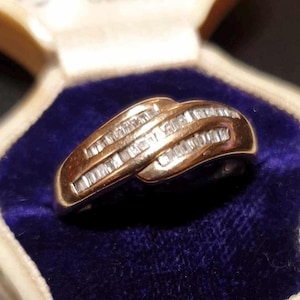 Puede incluir: Un anillo dorado con un diseño curvo, engastado con múltiples piedras preciosas pequeñas de corte rectangular. El anillo se exhibe sobre una superficie de terciopelo azul, mostrando sus intrincados detalles y su elegante estilo.