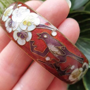 May include: A vintage enamel bracelet with a red background, depicting a purple bird on a branch and white floral details. The bracelet has a gold-coloured metal trim. The bracelet is held in a hand.
