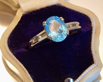 May include: A silver ring with a light blue oval gemstone and small clear gemstones set in the band. The ring is in a blue velvet ring box.