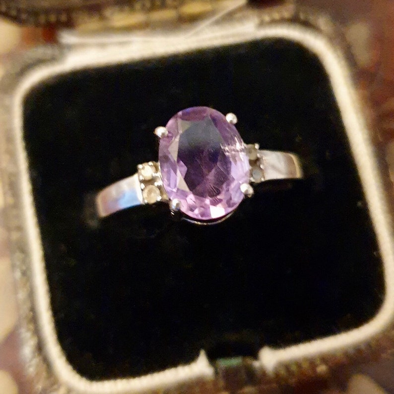 May include: A silver ring with a large oval amethyst gemstone and smaller accent stones. The ring is in a black velvet ring box.