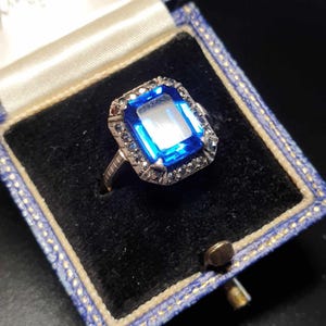 May include: A vintage ring featuring a large blue gemstone surrounded by smaller sparkling stones. The ring is set in silver and has a delicate design.