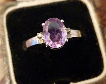 May include: A silver ring with a large oval amethyst gemstone and smaller accent stones. The ring is in a black velvet ring box.
