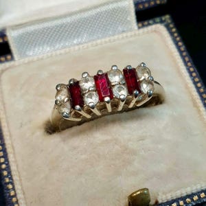 May include: A silver ring featuring a row of alternating red and clear gemstones. The ring is presented in a vintage jewellery box with a blue and white patterned interior.