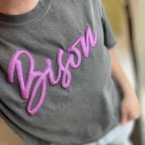 Könnte beinhalten: Dunkelgraues T-Shirt mit dem Wort "Bison" in glitzernder, kursiver, magenta Schrift auf der Brust. Das Shirt hat ein leicht abgenutztes Aussehen.
