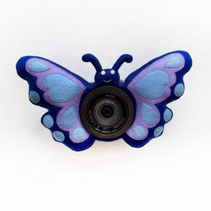 Op de afbeelding: Een blauwe en paarse vilten vlinder camerahoes. De hoes heeft een lachend gezicht en antennes. De cameralens is zichtbaar in het midden van de vlinder. De vleugels hebben een gelaagd ontwerp met lichtblauwe en paarse accenten.
