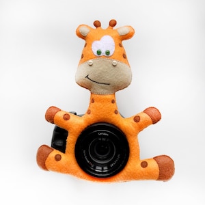 May include: A plush giraffe camera lens cover in orange and brown with a white face and green eyes. The giraffe is sitting on a camera lens with the text "Carl Zeiss" printed on the lens.
