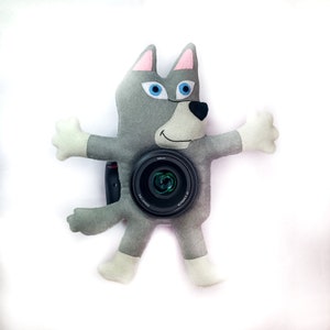 May include: A gray wolf plush toy with blue eyes and a pink nose is holding a black camera lens in front of its body.