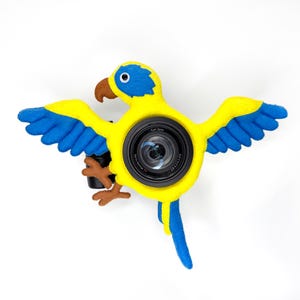 May include: A felt parrot camera cover featuring a bright yellow body, blue wings, and a brown beak. The parrot is designed with a central circular opening that fits around a black camera lens, which displays the text Carl Zeiss Vario-Sonnar.