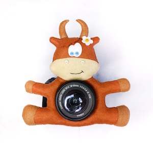 May include: A brown felt cow with a white flower on its head is holding a black Canon camera lens with the text "Canon Zoom Lens EF 5 18-55mm 1:3.5-5.6 IS STM 55mm" printed on it.
