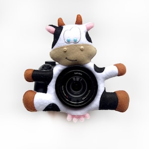 May include: A black and white cow-shaped camera lens cover with brown ears and a pink tail. The cover is made of felt and has a cute, cartoonish design. The lens cover is attached to a camera lens with a black lens cap.