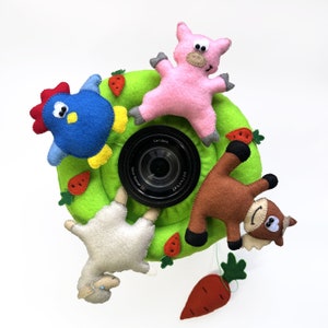 May include: A bright green felt ring featuring four plush farm animals: a blue chicken, a pink pig, a brown horse, and a white sheep. Small orange felt carrots are attached to the ring, which surrounds a black camera lens with Carl Zeiss text.