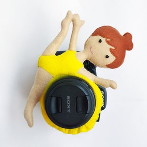May include: A handmade felt ballerina doll in a yellow leotard, posed over a black camera lens cap. The doll has brown hair and a smiling face. The lens cap is labeled "SONY".