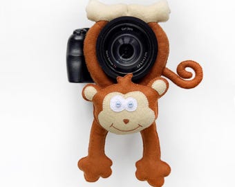 May include: A black camera with a brown felt monkey cover featuring a cream-colored face, two small button eyes, and a curled tail. The camera lens is visible through the center of the monkey's body, with the text Carl Zeiss Vario-Sonnar T* printed on it.