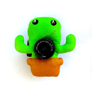 May include: A green felt cactus with a smiling face and black eyes holds a black Canon EF-S 18-55mm f/3.5-5.6 IS STM lens in front of it. The cactus is in a brown felt pot.