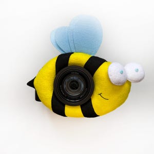 Könnte beinhalten: Ein Kameraobjektivdeckel in Form einer Hummel mit einem gelb-schwarz gestreiften Filzkörper, hellblauen Filzflügeln und zwei weißen Filzaugen mit kleinen blauen Pupillen. Das schwarze Kameraobjektiv ist in der Mitte des Hummelkörpers sichtbar.