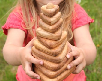 May include: A child holds a wooden stacking toy shaped like a pyramid with heart-shaped pieces. The toy is made of natural wood and has a light brown color.