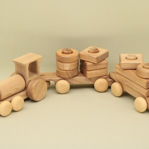 May include: A wooden toy train with three cars. The engine car has a large, round, wooden body. The middle car has a stack of wooden rings and the last car has a stack of wooden blocks. All the cars have wheels.