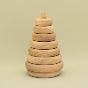 May include: A wooden stacking toy featuring seven graduated rings and a top peg. The natural wood rings are arranged in a pyramid shape, with the largest ring at the base. The toy is designed for children.
