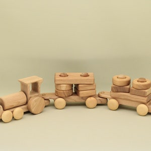 Op de afbeelding: Een houten speelgoedtrein met drie wagons. De locomotief heeft een groot, rond houten wiel. De middelste wagon heeft een stapel houten blokken en de laatste wagon heeft een stapel houten ringen. De trein is gemaakt van natuurlijk hout.