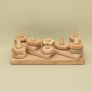 May include: Wooden stacking toy with various shapes and sizes, including circles, stars, and flowers. The pieces are arranged on a wooden base.