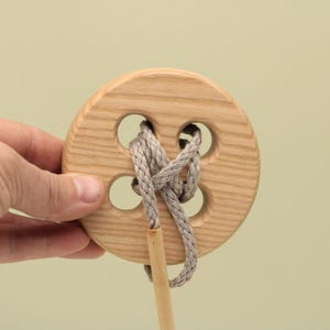 Op de afbeelding: Een houten knoop-puzzel met een lichtbruine afwerking. De knoop heeft vier gaten en een grijs touw dat erdoorheen is geregen. Een kleine houten deuvel is aan het touw bevestigd. De knoop wordt door een hand vastgehouden.