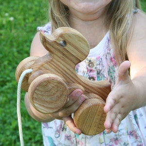 May include: A wooden pull toy shaped like a duck with four wheels. The toy is made of natural wood and has a light brown colour. A child is holding the toy in their hands.