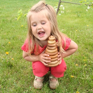 May include: A young girl wearing a red and white polka dot dress is playing with a wooden stacking toy. The toy is made of rings of different sizes that can be stacked on top of each other. The girl is smiling and looks happy to be playing with the toy.
