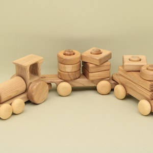 May include: A wooden toy train with three carriages. The engine carriage has a large, round, wooden body. The middle carriage has a stack of wooden rings and the last carriage has a stack of wooden blocks. All the carriages have wheels.