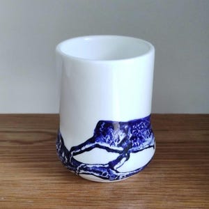 May include: A white ceramic tumbler with a cylindrical shape. The bottom portion features a striking abstract design in deep blue, resembling a landscape or geological formation. The cup sits on a wooden surface.