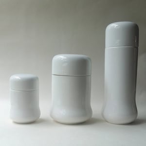 Può includere: Tre contenitori in ceramica bianca di diverse dimensioni. I contenitori sono cilindrici con parte superiore e inferiore arrotondate. I contenitori sono tutti della stessa tonalità di bianco.