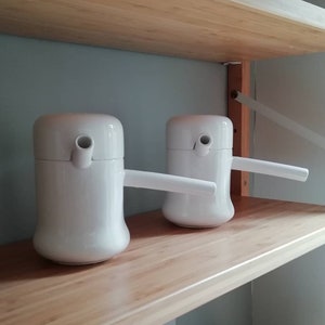 May include: Two white ceramic pitchers with a spout and a handle on a wooden shelf. The pitchers have a rounded body and a slightly flared top.