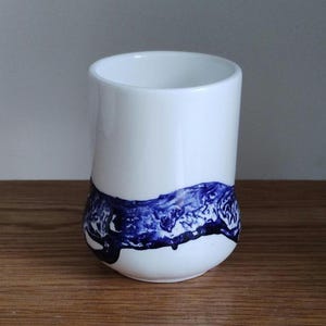 May include: A white ceramic tumbler with a rounded base and a cylindrical shape. The bottom third of the tumbler features a dark blue, abstract design. The tumbler is set on a wooden surface.