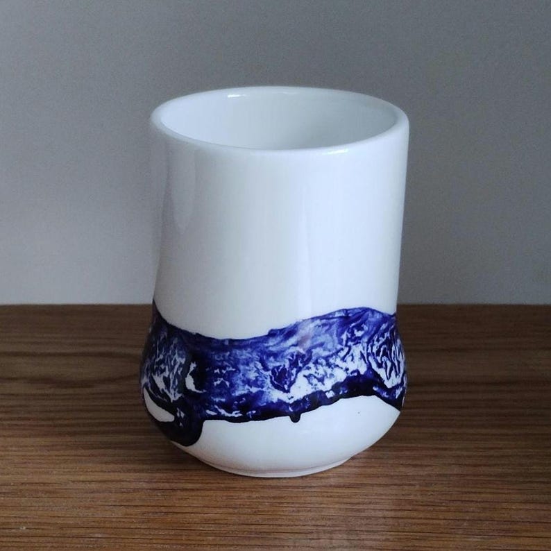 May include: A white ceramic tumbler with a rounded base and a cylindrical shape. The bottom third of the tumbler features a dark blue, abstract design. The tumbler is set on a wooden surface.