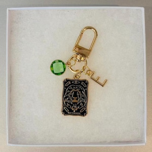 May include: A gold-toned keychain featuring a rectangular black enamel charm with a lion illustration and the text "THE STRENGTH", a small green faceted crystal bead, and a gold-toned letter "E" charm attached to a lobster claw clasp.
