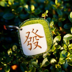 May include: A green and white mahjong tile keychain with the Chinese character "發" (pronounced "fa") in gold lettering. The tile is surrounded by a green glitter border.