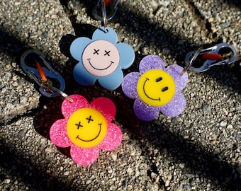 May include: Three flower-shaped keychains in pink, blue, and purple, each with a smiling face design. The pink flower has a yellow center, the blue has a light pink center, and the purple has a yellow center. Each keychain has a silver clasp.