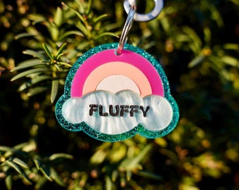 Può includere: Un arcobaleno rosa e turchese con una nuvola bianca su cui è scritto "FLUFFY". L'etichetta è in acrilico e ha una finitura glitter.