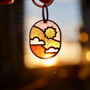Op de afbeelding: Een ovaalvormige sleutelhanger met een landschapsontwerp. Het ontwerp toont een gele zon, witte wolken en oranje en gele heuvels. De sleutelhanger wordt omhoog gehouden tegen een wazige achtergrond van een zonsondergang.