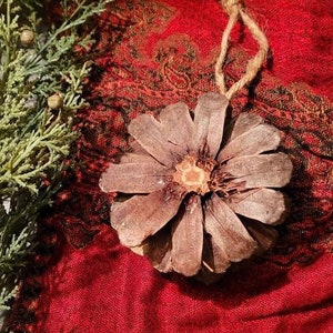 以下が含まれることがあります： 赤い金色の模様のフリンジ付きの生地の上に置かれた、素朴な麻ひものハンガーが付いた茶色の松ぼっくりオーナメント