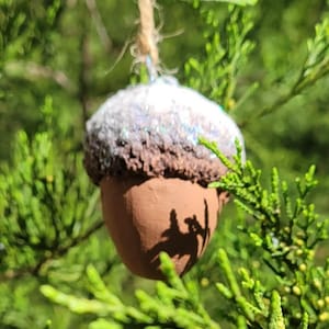 May include: A brown and silver acorn ornament with glitter on the top. The ornament is hanging from a tree branch.
