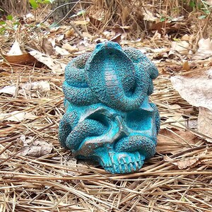 May include: A teal-colored skull sculpture with a cobra coiled around it. The skull has detailed features, and the snake's scales are textured. The sculpture is set against a backdrop of dry leaves and foliage.
