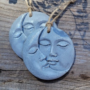 May include: Two gray, ceramic moon ornaments with faces and a crescent moon design. The ornaments are hanging from twine.