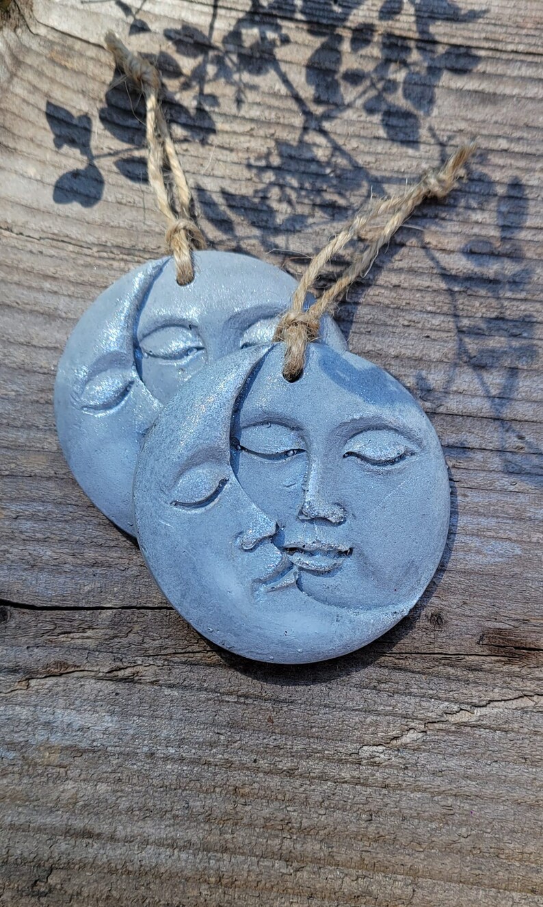 May include: Two gray, ceramic moon ornaments with faces and a crescent moon design. The ornaments are hanging from twine.