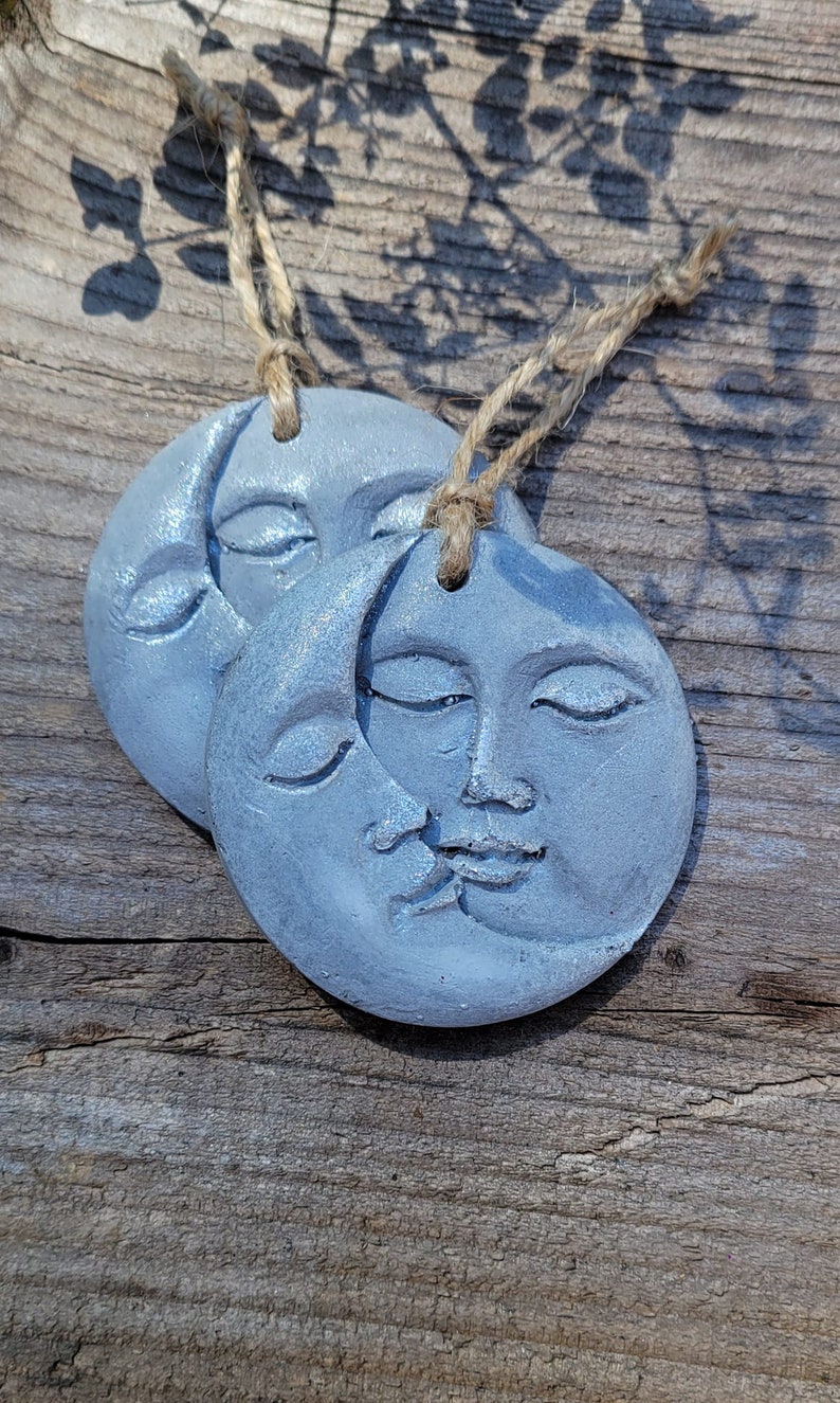 May include: Two gray, ceramic moon ornaments with faces and a crescent moon design. The ornaments are hanging from twine.